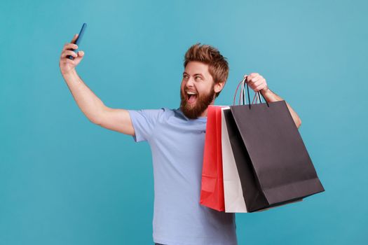 Portrait of positive handsome young adult bearded man holding shopping bags and talking selfie on smartphone, satisfied with purchase. Indoor studio shot isolated on blue background.