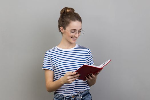 Portrait of satisfied smiling woman wearing striped T-shirt and glasses holding book, enjoys reading, standing absorbed with exciting plot. Indoor studio shot isolated on gray background.