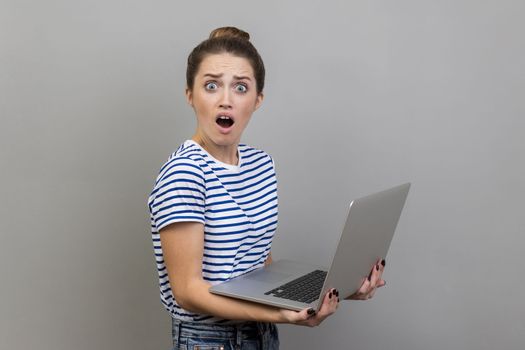 Side view of shocked woman wearing striped T-shirt working on laptop with surprised expression, freelance work, looking at camera with open mouth. Indoor studio shot isolated on gray background.