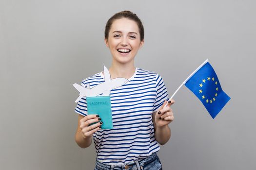 Portrait of woman wearing striped T-shirt standing with Europe union flag and passport with paper plane, traveling to european country. Indoor studio shot isolated on gray background.