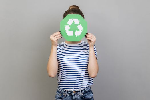 Portrait of anonymous unknown woman wearing striped T-shirt covering her face with green recycling sign, environmental problems. Indoor studio shot isolated on gray background.