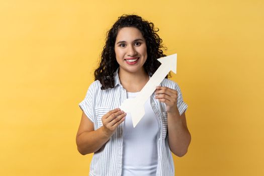 Portrait of smiling satisfied woman with dark wavy hair pointing direction with arrow aside, looking at camera with positive expression. Indoor studio shot isolated on yellow background.