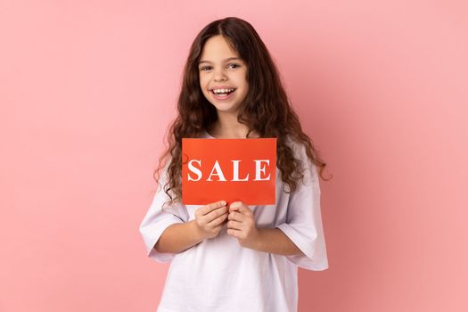 Portrait of happy delighted little girl wearing white T-shirt holding card with sale inscription, looking at camera with toothy smile. Indoor studio shot isolated on pink background.