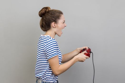 Side view portrait of woman wearing striped T-shirt holding gamepad, winning video games competition, screaming happy expression. Indoor studio shot isolated on gray background.