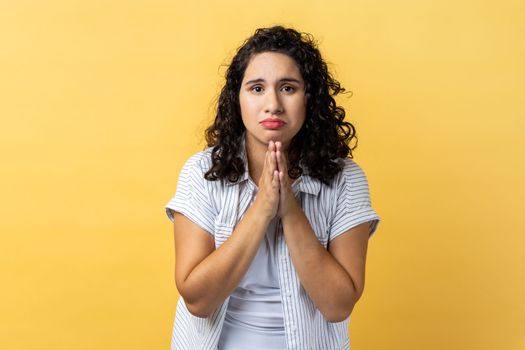 Portrait of woman with dark wavy hair holding hands in prayer, looking with imploring pleading expression, begging help, asking forgiveness. Indoor studio shot isolated on yellow background.