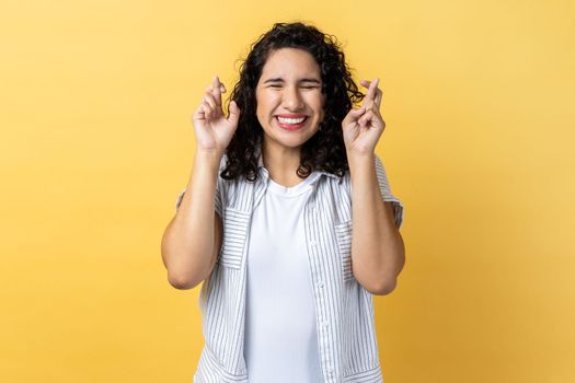 Portrait of hopeful adorable woman with dark wavy hair standing with crossed fingers, wishes her dream comes true, keeps eyes closed. Indoor studio shot isolated on yellow background.