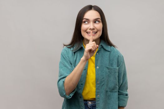 Positive woman shushing with silence gesture, holding finger on lips and smiling to camera, asking to keep secret, wearing casual style jacket. Indoor studio shot isolated on gray background.