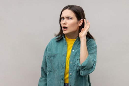 Portrait of curious woman holding hand near ear trying to hear secrets and find out gossips, listen to private whispers, wearing casual style jacket. Indoor studio shot isolated on gray background.