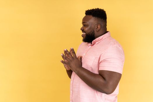 Side view portrait of cunning bearded man wearing pink shirt clasping hands and planning evil tricky prank or scheming, cheating with sly smile. Indoor studio shot isolated on yellow background.