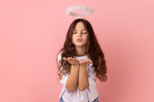 Portrait of little girl wearing white T-shirt with halo on head sending air kiss on camera, flirting, valentines day, keeps eyes closed. Indoor studio shot isolated on pink background.