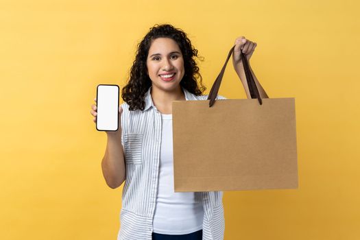 Portrait of smiling happy woman with dark wavy hair holding paper shopping bag and smart phone with empty display for advertisement. Indoor studio shot isolated on yellow background.
