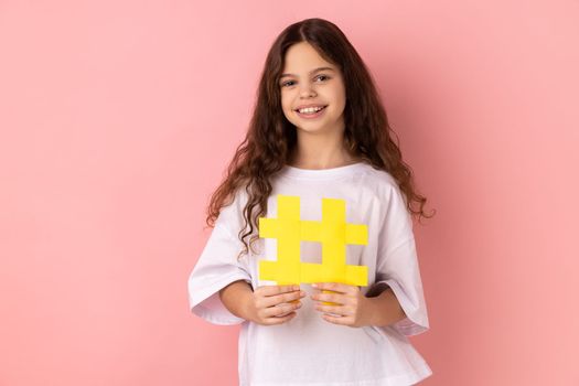Portrait of adorable little girl wearing white T-shirt holding hashtag symbol and looking at camera with toothy smile, showing hash sign. Indoor studio shot isolated on pink background.