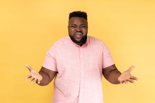 Portrait of unhappy stressed man wearing pink shirt standing raised arms and asking, expressing negative emotions, screaming and frowning face. Indoor studio shot isolated on yellow background.