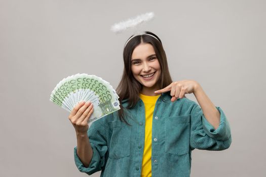 Cheerful dark haired woman with halo on head pointing finger on bunch of euro banknotes and smiling, profit, wearing casual style jacket. Indoor studio shot isolated on gray background.