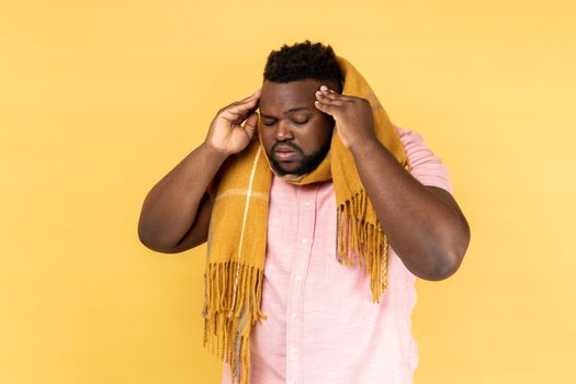 Portrait of unhealthy man wearing pink shirt standing wrapped in scarf, having terrible headache, massaging temples, flu symptoms. Indoor studio shot isolated on yellow background.