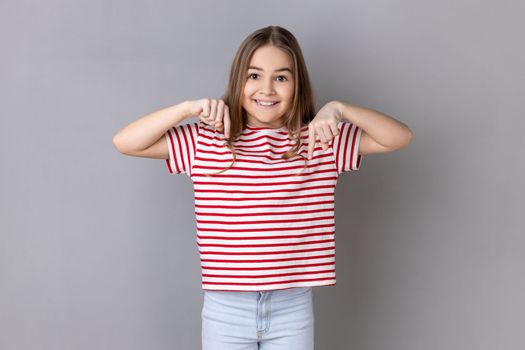 Look, advertise below. Portrait of little girl wearing striped T-shirt pointing down place for commercial idea, looking at camera with toothy smile. Indoor studio shot isolated on gray background.
