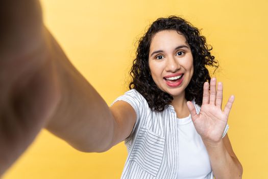 Portrait of smiling friendly woman with dark wavy hair making point of view photo, waving hand, saying hello or good bye, POV. Indoor studio shot isolated on yellow background.