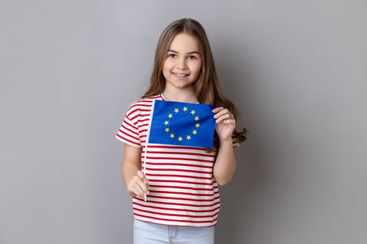 European Union flag. Portrait of smiling adorable cute little girl wearing striped T-shirt holding Europe flag, looking at camera. Indoor studio shot isolated on gray background.