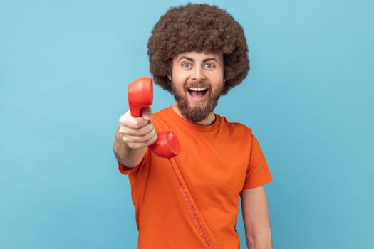 Portrait of man with Afro hairstyle wearing orange T-shirt holding and showing retro phone handset to camera, asking to answer phone. Indoor studio shot isolated on blue background.