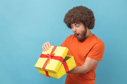 Portrait of man with Afro hairstyle wearing orange T-shirt unpacking present, looking inside box with shocked and scared expression, birthday gist. Indoor studio shot isolated on blue background.