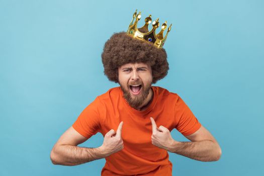 Portrait of crazy man with Afro hairstyle in golden crown and pointing himself with excite expression, looking with arrogance, declaring his authority. Indoor studio shot isolated on blue background.
