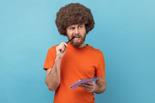 Portrait of man with Afro hairstyle in orange T-shirt standing thoughtful biting pencil, pondering idea to write into notebook, taking notes in paper. Indoor studio shot isolated on blue background.
