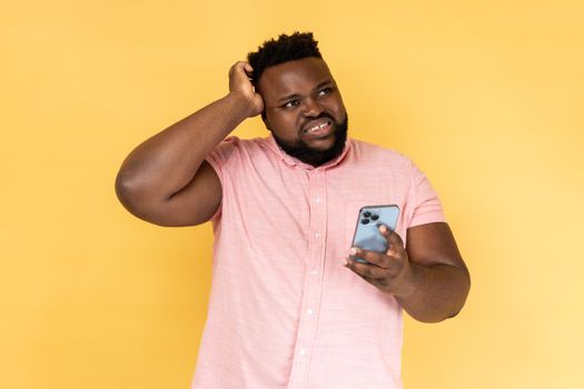 Portrait of pensive thoughtful bearded man wearing pink shirt standing with mobile phone, touching head, having doubt suspicion feeling. Indoor studio shot isolated on yellow background.