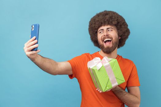 Portrait of happy joyful man with Afro hairstyle wearing orange T-shirt having video call with present box, boasting his gift to followers. Indoor studio shot isolated on blue background.