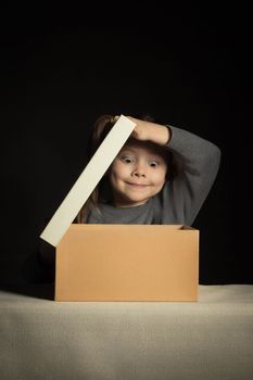 Emotional happy and surprised little girl opens a long-awaited package with a gift, copy space on cardboard box, vertical.