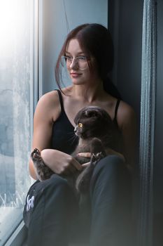 Attractive teenage girl sits alone on the windowsill together with a pet python with a cat and looks out the window. High quality photo