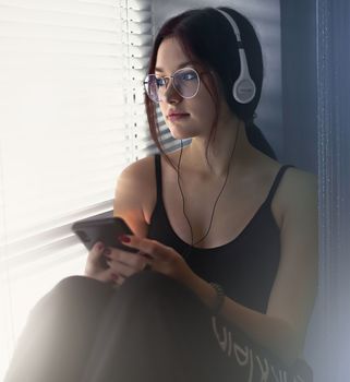 Attractive teenage girl sitting on the windowsill alone listening to music and looking out the window. High quality photo