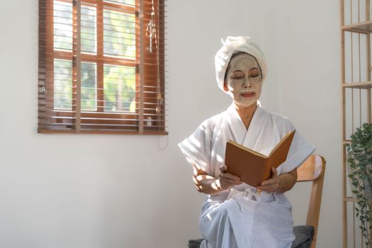 Beautiful mature Asian lady in bathrobe with facial moisturizing or anti aging clay mask on her face is reading a book in living room