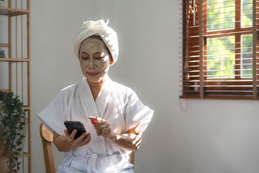 Beautiful mature Asian lady in bathrobe with facial moisturizing or anti aging clay mask on her face is using her mobile phone