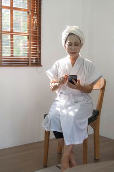 Beautiful mature Asian lady in bathrobe with facial moisturizing or anti aging clay mask on her face is using her mobile phone