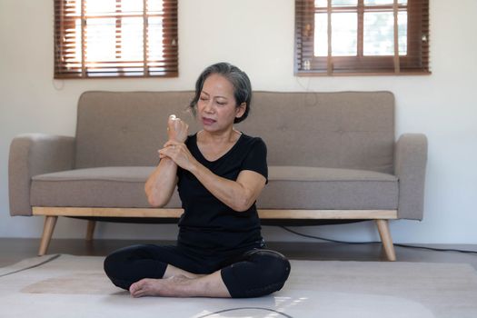 60s aged Asian woman massaging her wrist, feeling pain and swelling in the joints, injured hand during yoga practice at home...