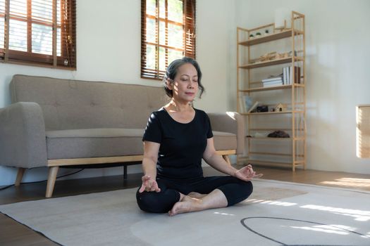 Calm and relaxed 60s retired Asian woman in gym clothes is meditating in her living room, eyes closed, practicing lotus yoga pose at home...