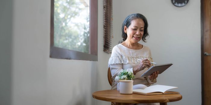 60s aged Asian woman in casual clothes sitting by the window in her living room and using portable digital tablet