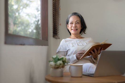 Relaxed and calm 60s retired Asian woman looking out the window, daydreaming while reading a book in her home living room. lifestyle concept..