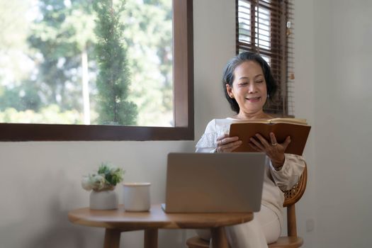 Relaxed and calm 60s retired Asian woman looking out the window, daydreaming while reading a book in her home living room. lifestyle concept..