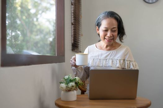 Portrait, Professional and successful aged Asian businesswoman working from home, sipping morning coffee while using laptop computer...