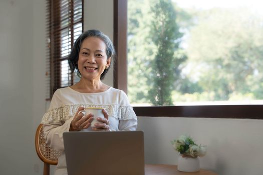 Portrait, Professional and successful aged Asian businesswoman working from home, sipping morning coffee while using laptop computer...