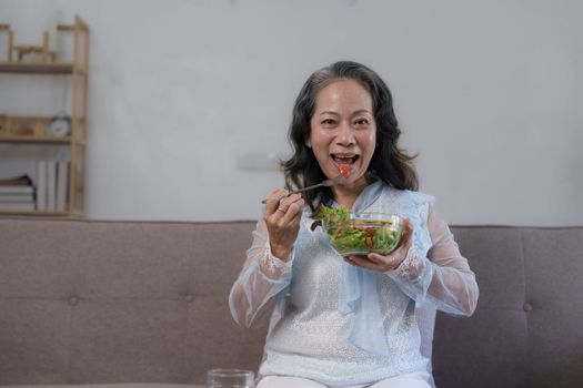 Portrait, Cheerful Asian 60s aged woman having breakfast in her living room, eating healthy salad vegetables mix bowl...