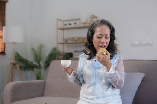 Beautiful and happy Asian 60s aged woman having breakfast, sipping a cup of tea and eating cookies in her living room...