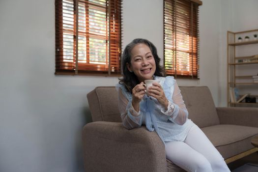 Portrait, Relaxed and calm 60s aged Asian woman sipping hot tea, having an afternoon tea in her living room. lifestyle concept..