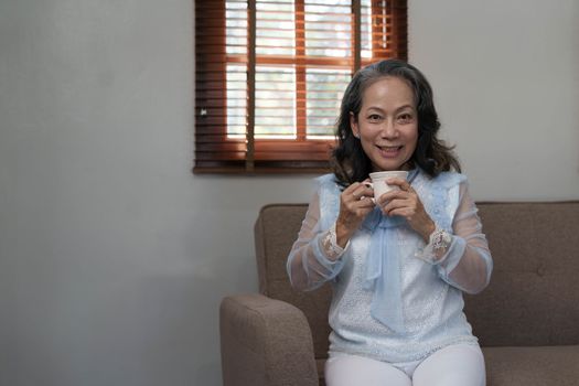 Portrait, Relaxed and calm 60s aged Asian woman sipping hot tea, having an afternoon tea in her living room. lifestyle concept..