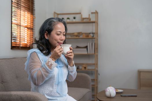 Portrait, Relaxed and calm 60s aged Asian woman sipping hot tea, having an afternoon tea in her living room. lifestyle concept..