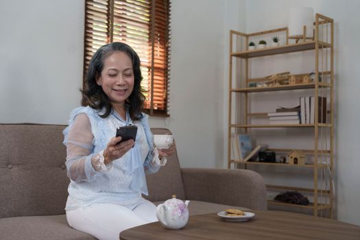 Cheerful 60s retired Asian woman using her mobile phone to chat with her grandchild while relaxing in her living room...