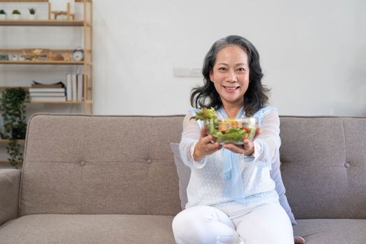 Portrait, Cheerful Asian 60s aged woman having breakfast in her living room, eating healthy salad vegetables mix bowl...