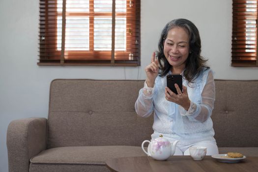 Cheerful 60s retired Asian woman using her mobile phone to chat with her grandchild while relaxing in her living room...