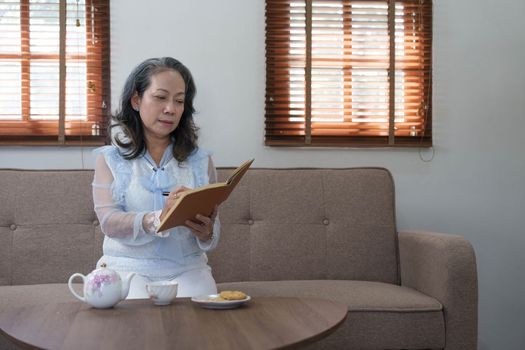 Portrait, Gorgeous 60s aged Asian woman in casual clothes eating cookies and writing her diary while relaxing in her living room..
